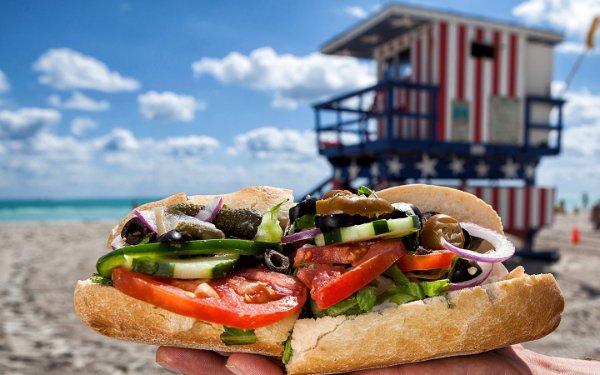 A close-up of a fresh sandwich with layers of vibrant ingredients, set against the backdrop of a sunny beach and lifeguard stand